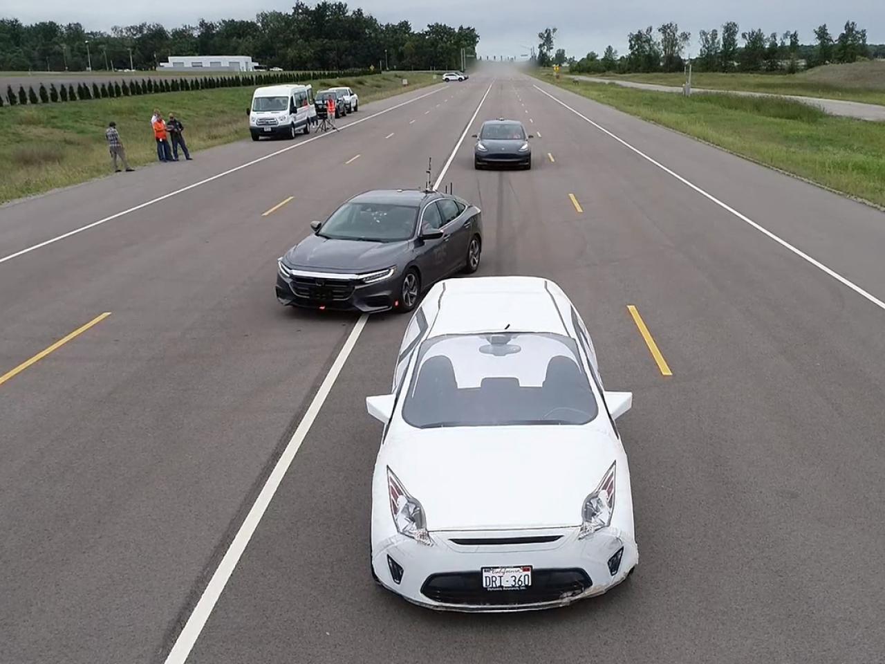 Three cars on a multi-lane road, with people standing near parked vehicles on the shoulder.